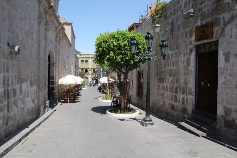 오래된 석조 건물 옆에 야외 테이블과 우산이 있는 자갈길. 나무와 가로등이 중앙에 있습니다.