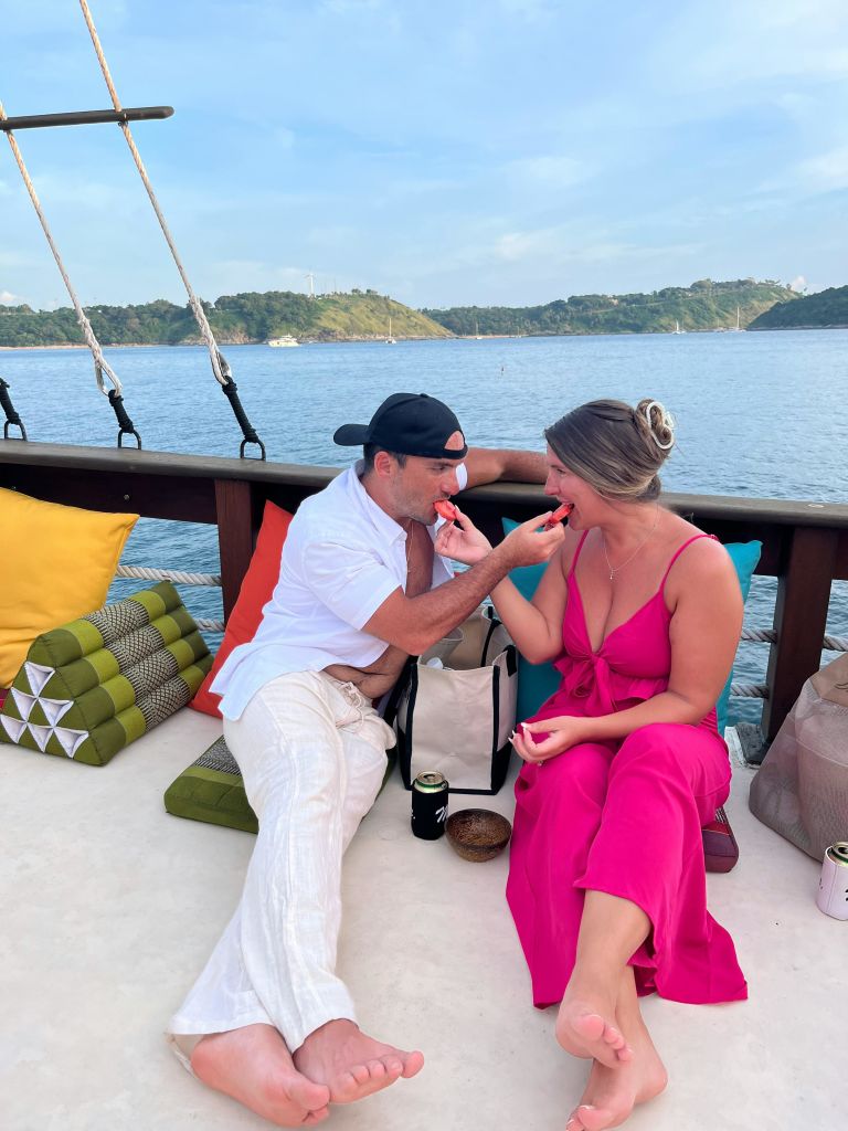 흰색 옷을 입은 남성과 분홍색 드레스를 입은 여성이 아름다운 물과 섬을 배경으로 보트에서 수박 조각을 먹고 있습니다.