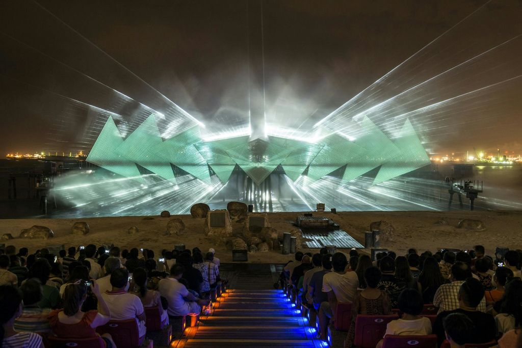 야외 공연장에 앉아 무대에서 녹색 기하학적 모양과 대들보로 빛이 투영되는 쇼를 감상하는 사람들.