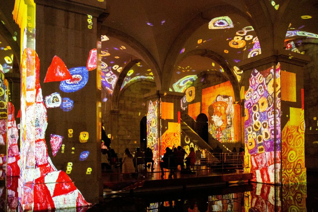 벽과 기둥에 화려한 추상 예술 투영이 있는 아치형 홀 내부의 사람들 (얼굴과 기하학적 모양 포함).