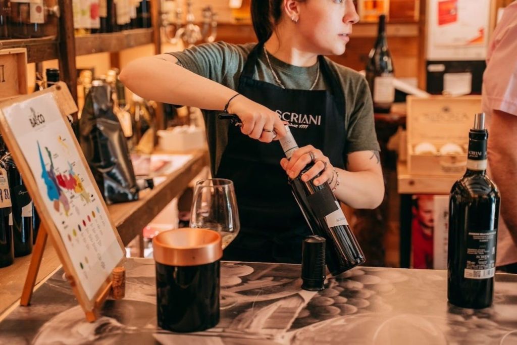 한 상점에서 앞치마를 두른 채 와인병을 열고 있는 사람이 앞에 놓인 테이블에 와인병과 유리잔을 놓고 있습니다.