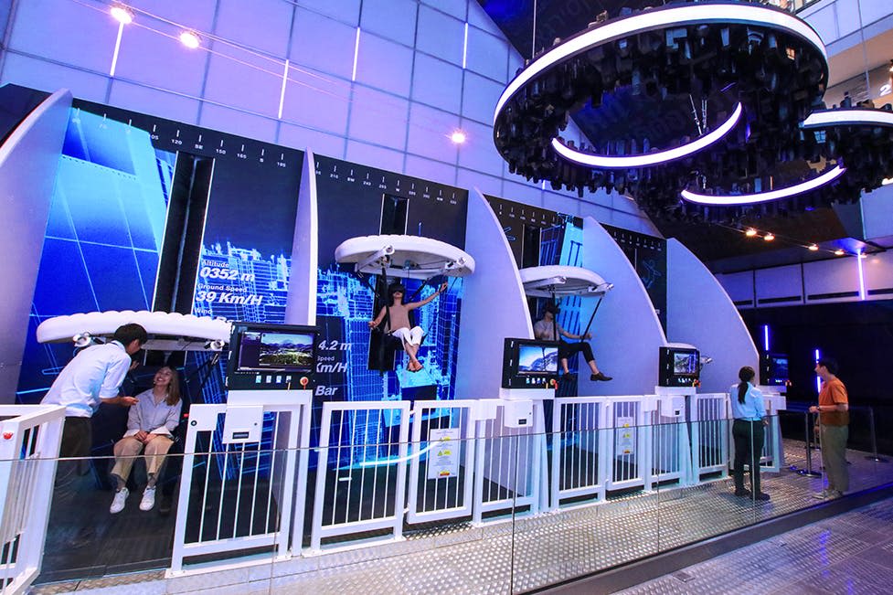 미래형 가상 현실을 사용하는 사람들은 파란색과 흰색 조명이 있는 실내 레크리에이션 공간에서 시뮬레이터를 탑니다.