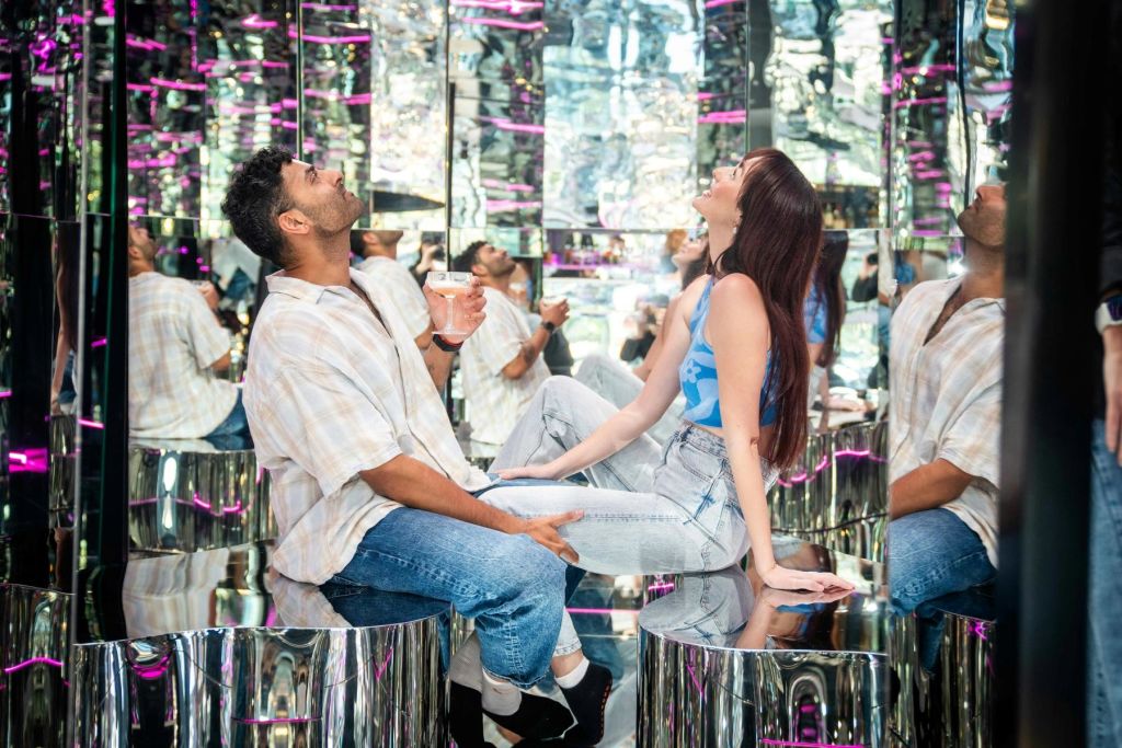 반사가 심한 방에서 거울로 된 의자에 앉아 음료를 들고 위를 바라보며 웃는 남녀.