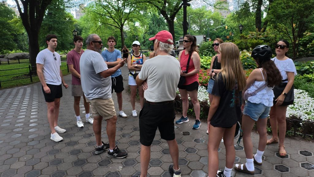 나무와 녹지로 둘러싸인 공원의 통로에 서 있는 한 무리의 사람들. 일부는 캐주얼한 여름 옷을 입고 있습니다.