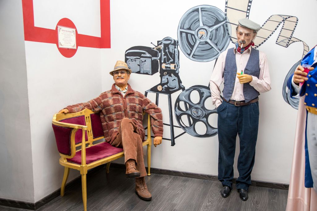 한 명은 착석하고 한 명은 서 있는 노인 복장을 한 두 마네킹이 필름 릴과 카메라가 그려진 벽 앞에서 포즈를 취하고 있습니다.