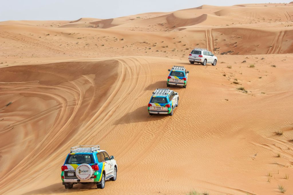 네 대의 SUV가 맑은 하늘 아래 모래사막 언덕을 달리고 있으며, 그 뒤로 타이어 트랙이 보입니다.