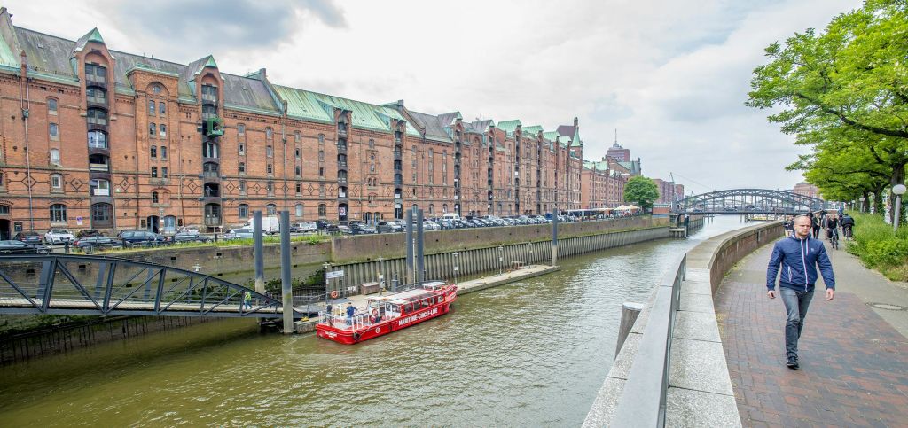 한 남자가 나무와 역사적 건물이 늘어선 강변 통로를 걷고 있습니다. 저 멀리 흐린 하늘 아래 다리가 있습니다.