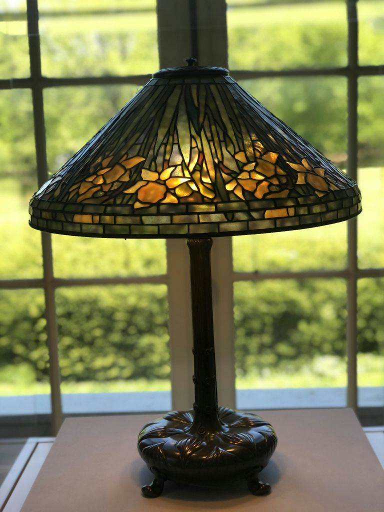 꽃무늬 디자인의 스테인드글라스 테이블 램프가 조명으로 켜져 있으며, 창문 앞에 녹지가 바깥에 보입니다.