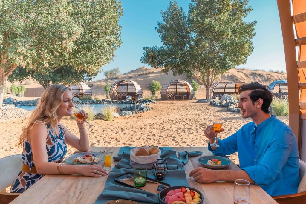 맑고 푸른 하늘 아래에서 경치 좋은 사막 글램핑 포드를 배경으로 야외에서 식사하는 파란색 셔츠를 입은 남성.