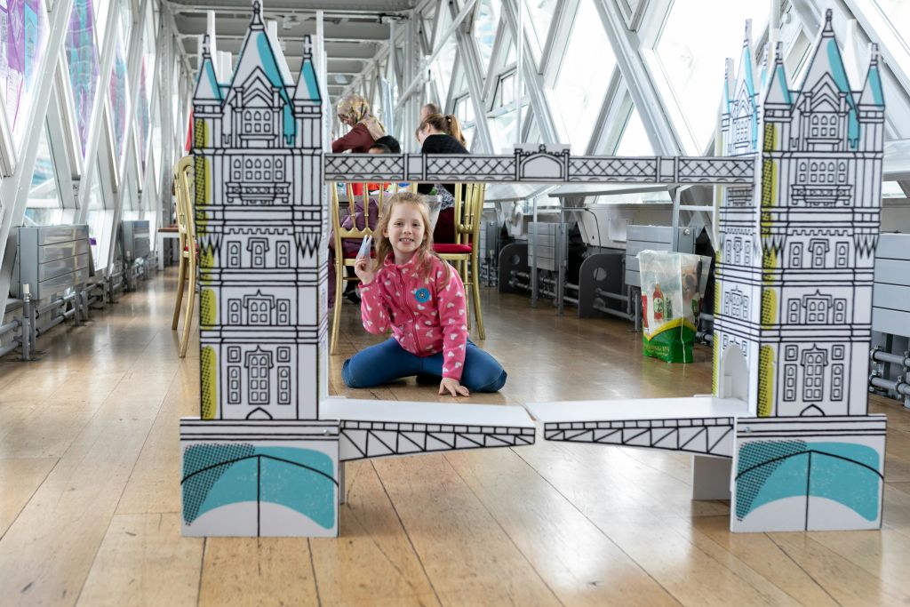 분홍색 재킷을 입은 어린이가 유리 벽으로 마감된 실내 공간에서 타워 브리지의 골판지 모형 옆에 평화 표지판을 들고 포즈를 취하고 있습니다.