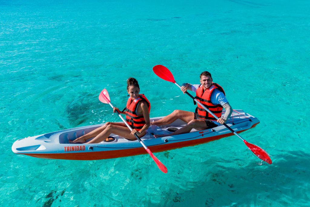 주황색 구명 조끼를 입은 두 사람이 청록색의 맑은 물 위에서 카약을 타고 빨간색 패들을 들고 카메라를 향해 미소를 짓고 있습니다.