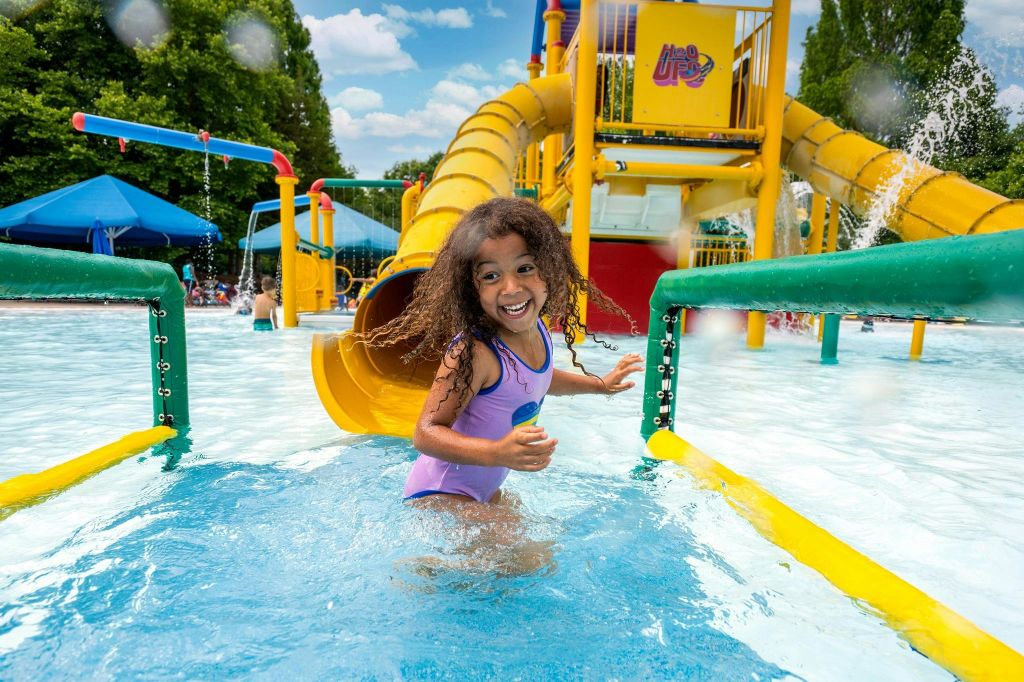 미끄럼틀과 물방울 구조물이 있는 워터파크에서 수영복을 입은 미소 짓는 어린이가 얕은 물속을 걷고 있습니다.