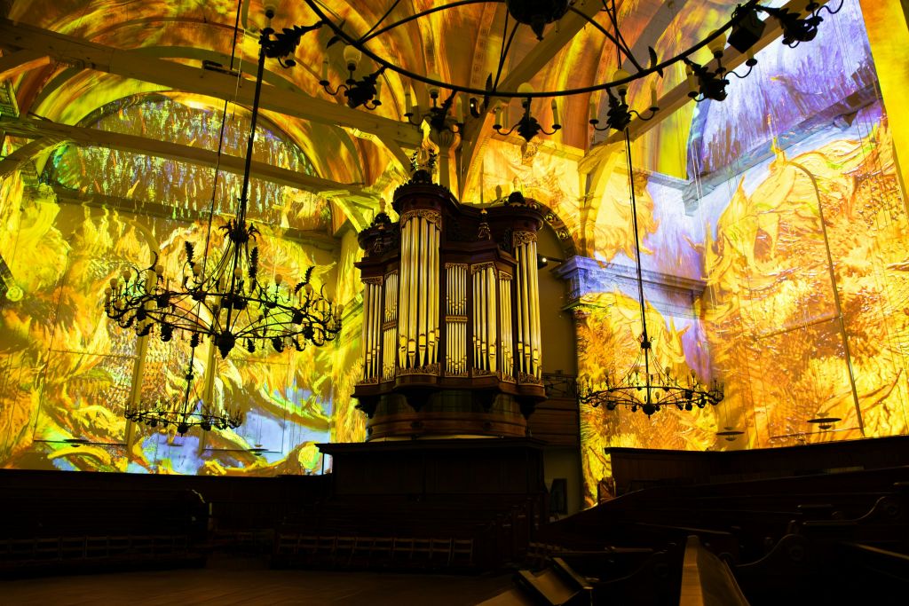 조명이 켜진 오르간이 있는 웅장한 교회 내부. 화려하고 추상적인 조명 투영이 벽과 천장을 뒤덮습니다.