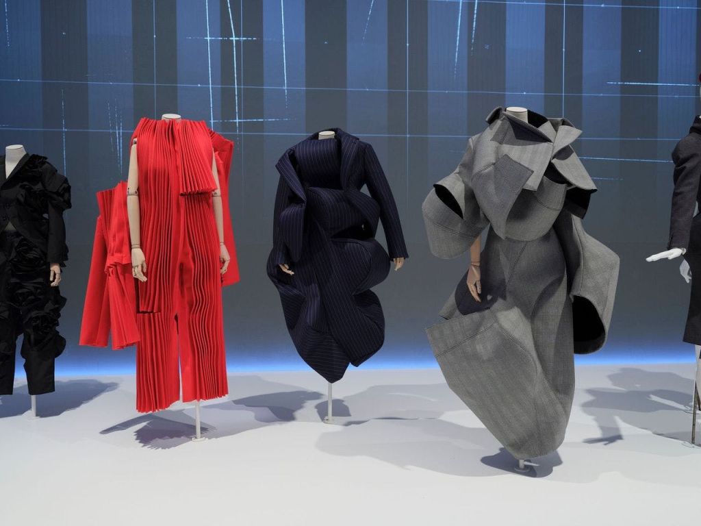 파란색 격자를 배경으로 빨간색 주름, 파란색 조각, 회색 레이어드 디자인의 아방가르드한 의상을 보여주는 세 개의 마네킹.