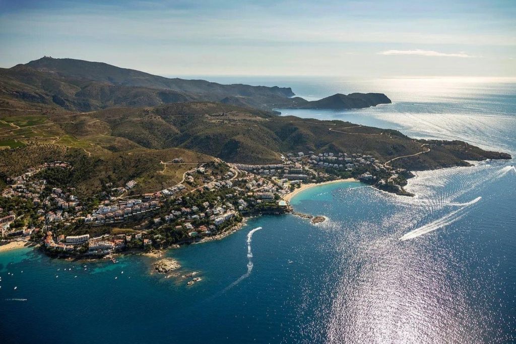맑은 하늘 아래 푸른 바다로 둘러싸인 만과 언덕을 배경으로 주택이 있는 해안 마을입니다.