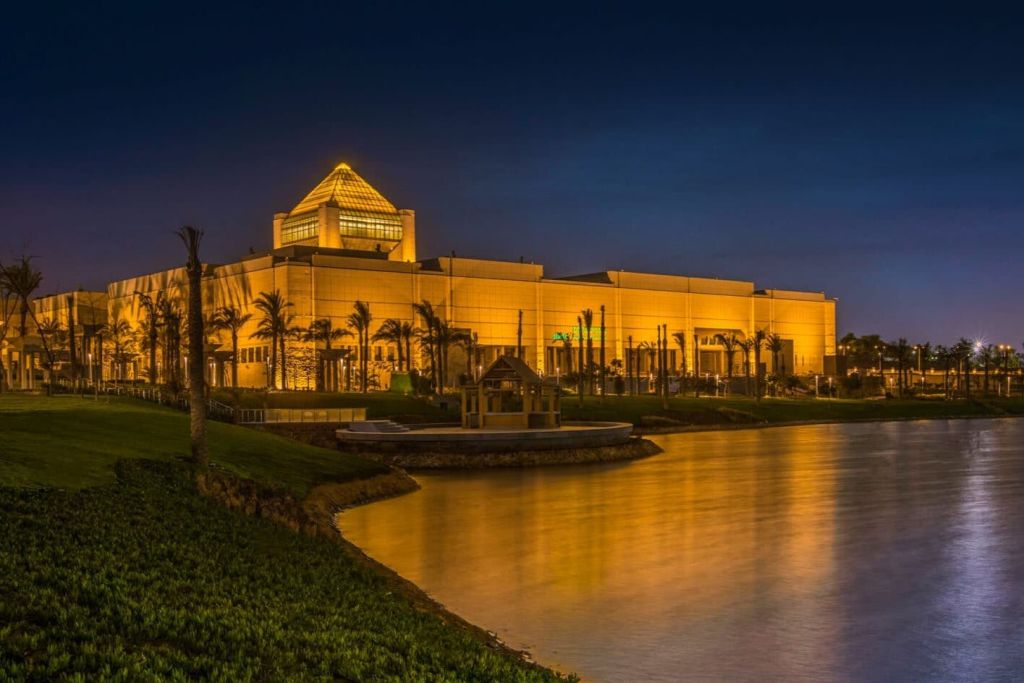 밤에는 깊고 푸른 하늘 아래 야자수와 물로 둘러싸인 유리 피라미드 지붕이 있는 조명 건물.