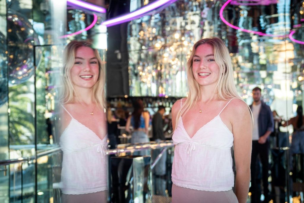 하얀 상의를 입은 여성이 반사되는 표면, 화려한 조명, 흐릿한 배경의 방에서 미소를 짓고 있습니다.