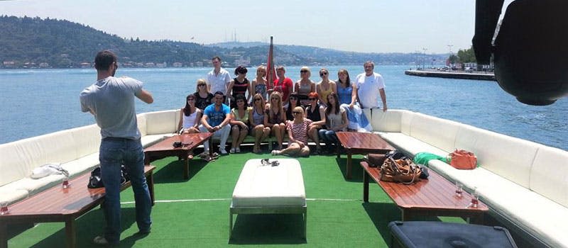 물과 먼 언덕을 배경으로 흰색 좌석과 녹색 카펫이 깔린 보트에서 포즈를 취하고 있는 한 무리의 사람들.