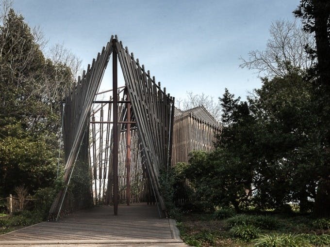 맑은 하늘 아래 나무와 녹음으로 둘러싸인 삼각형의 입구가 있는 목조 구조물. 내부로 이어지는 통로.