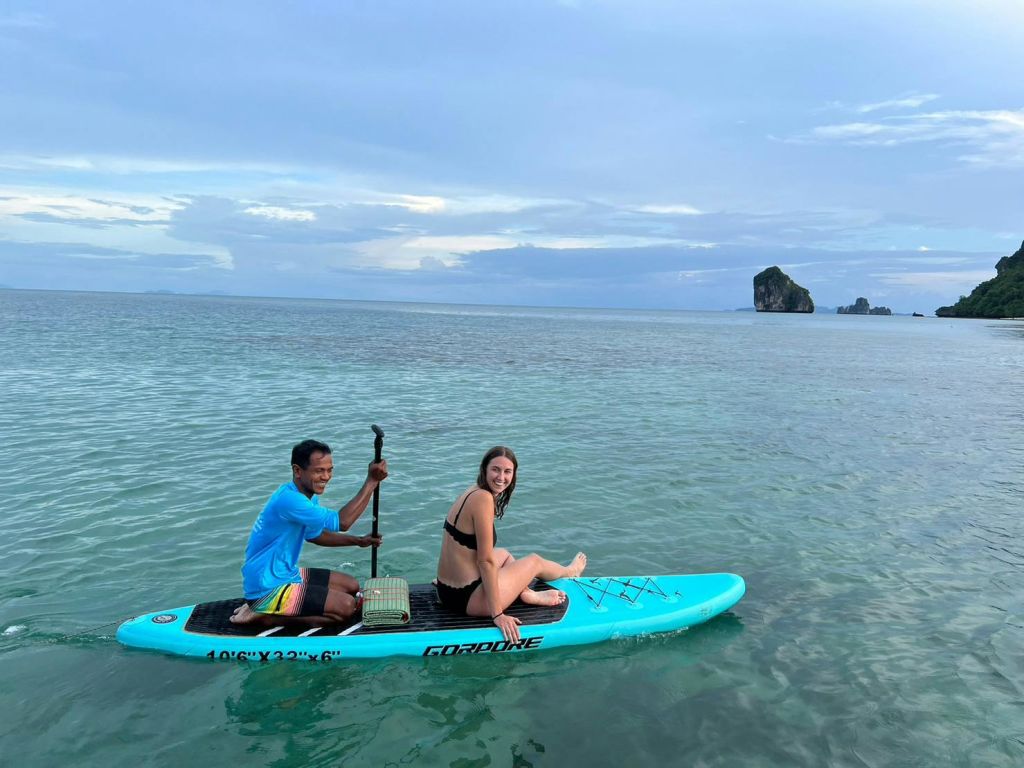 잔잔한 바닷물에서 청록색 스탠드업 패들보드에 앉아 웃고 있는 두 사람. 배경에는 작은 섬이 보입니다.