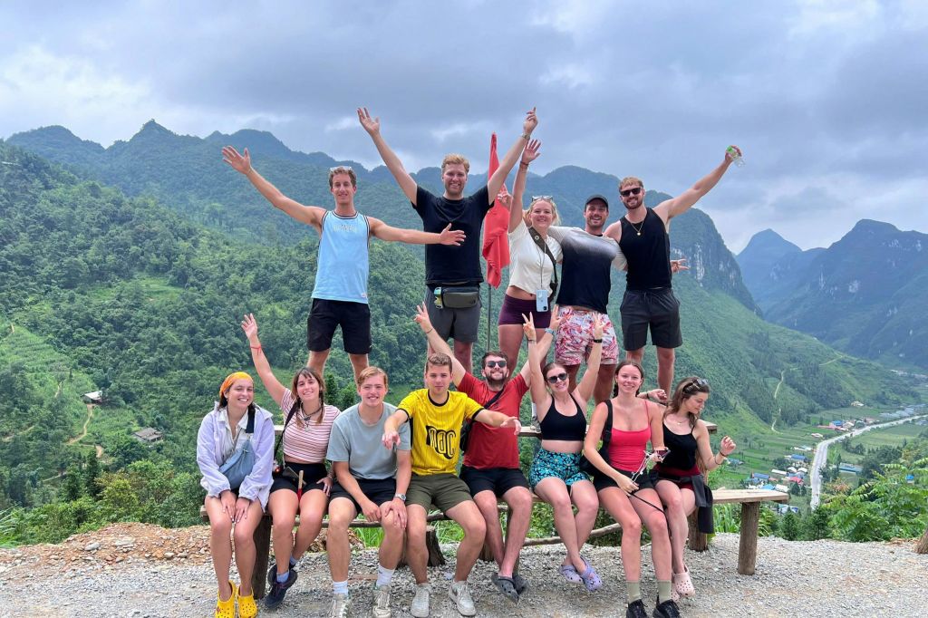 14명의 일행이 흐린 하늘 아래 산이 펼쳐진 풍경 앞에서 두 팔을 번쩍 들고 미소를 짓고 포즈를 취하고 있습니다.