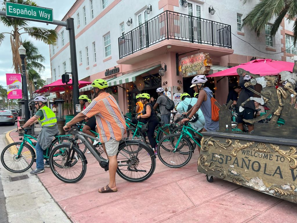 자전거를 탄 사람들은 발코니, 상점 표지판, 분홍색 파라솔이 있는 파스텔 색상의 건물 앞에 모였습니다.