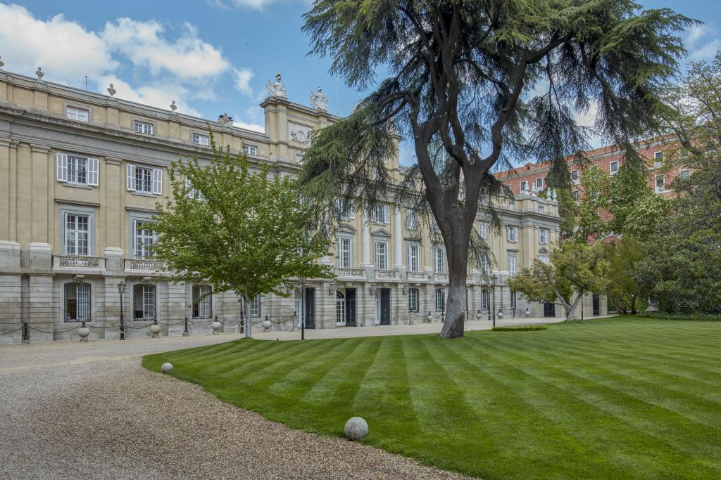 역사적인 복층 건물 앞에 자갈길, 커다란 나무, 잘 정비된 잔디가 있는 잘 가꾸어진 정원.
