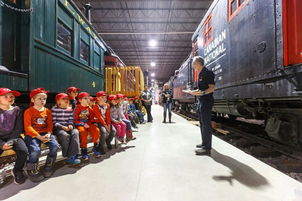 빨간 모자를 쓴 한 무리의 어린이들이 기차 박물관 안 벤치에 앉아 있으며, 양쪽에 빈티지 기차 차량이 있습니다.