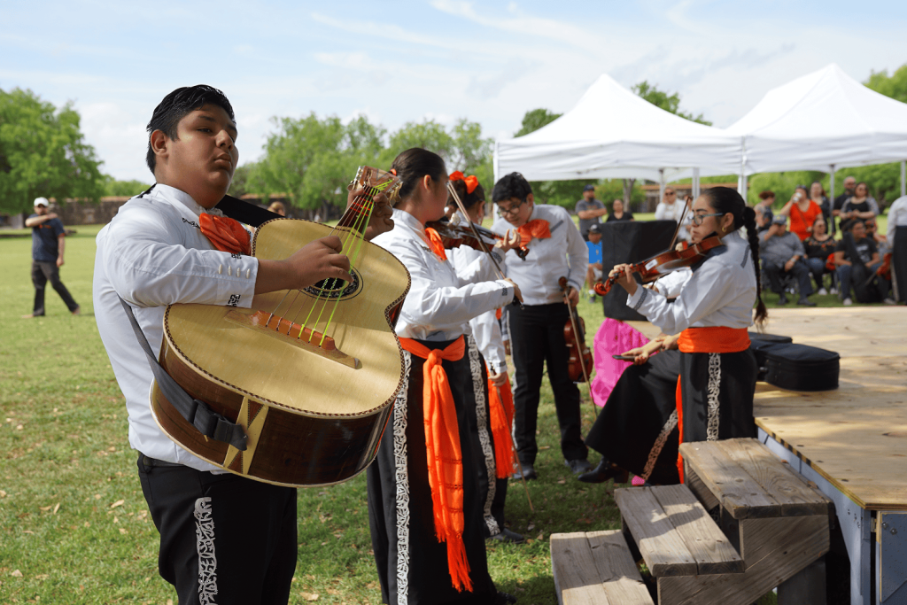 주황색 띠가 달린 흰색 의상을 입은 뮤지션이 야외에서 대형 기타, 바이올린 및 기타 악기를 연주합니다.