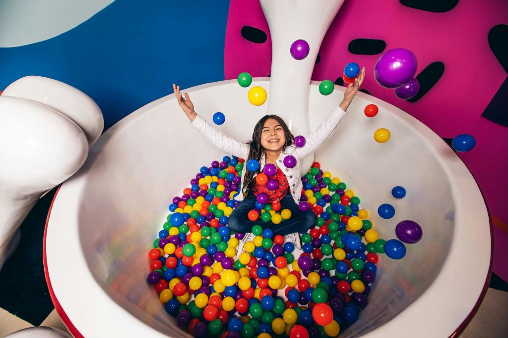 웃는 소녀는 장난기 가득한 환경에서 비슷한 공이 가득한 커다란 그릇에 앉아 화려한 공을 공중에 던집니다.