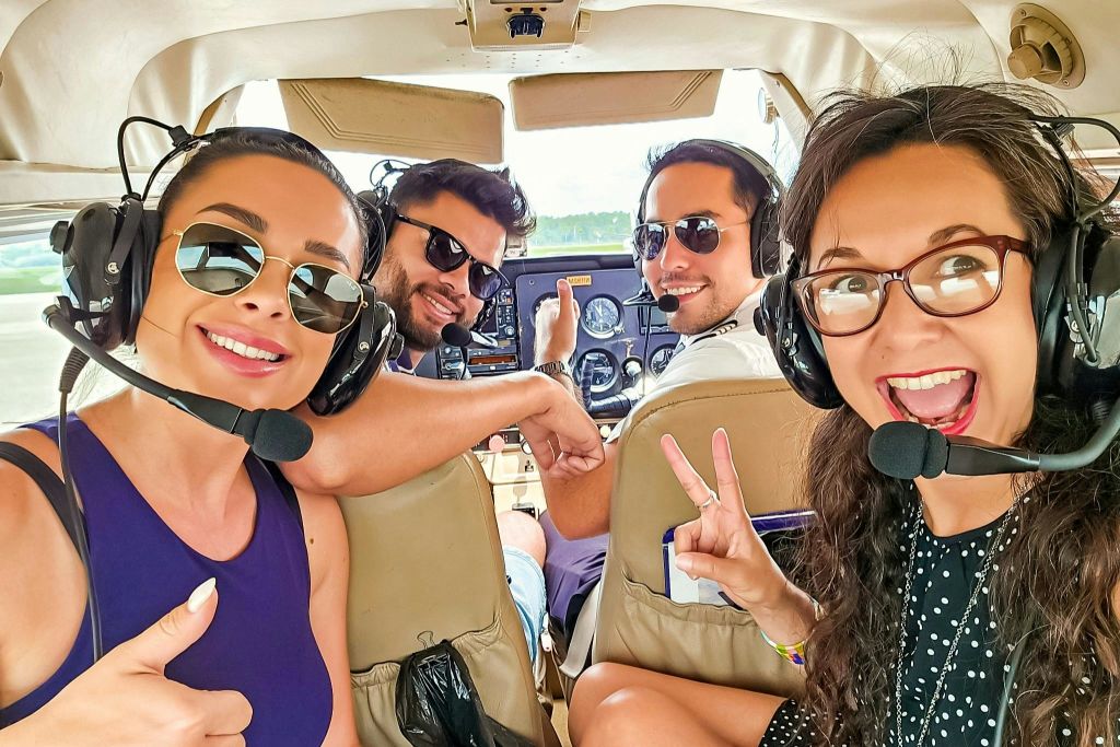 작은 비행기 조종실 안에 헤드셋을 착용하고 웃고 있는 네 명의 사람들. 두 명은 앞쪽에, 두 명은 뒤쪽에 앉아 평화의 표지판을 만들고 있습니다.