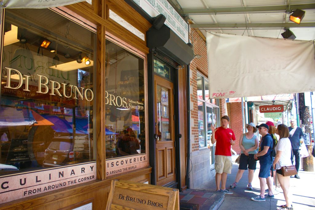 한 무리의 사람들이 벽돌 상점 앞에 서서 어닝 아래에 "Di Bruno Bros" 라고 적힌 표지판을 들고 투어 가이드의 말을 듣고 있습니다.