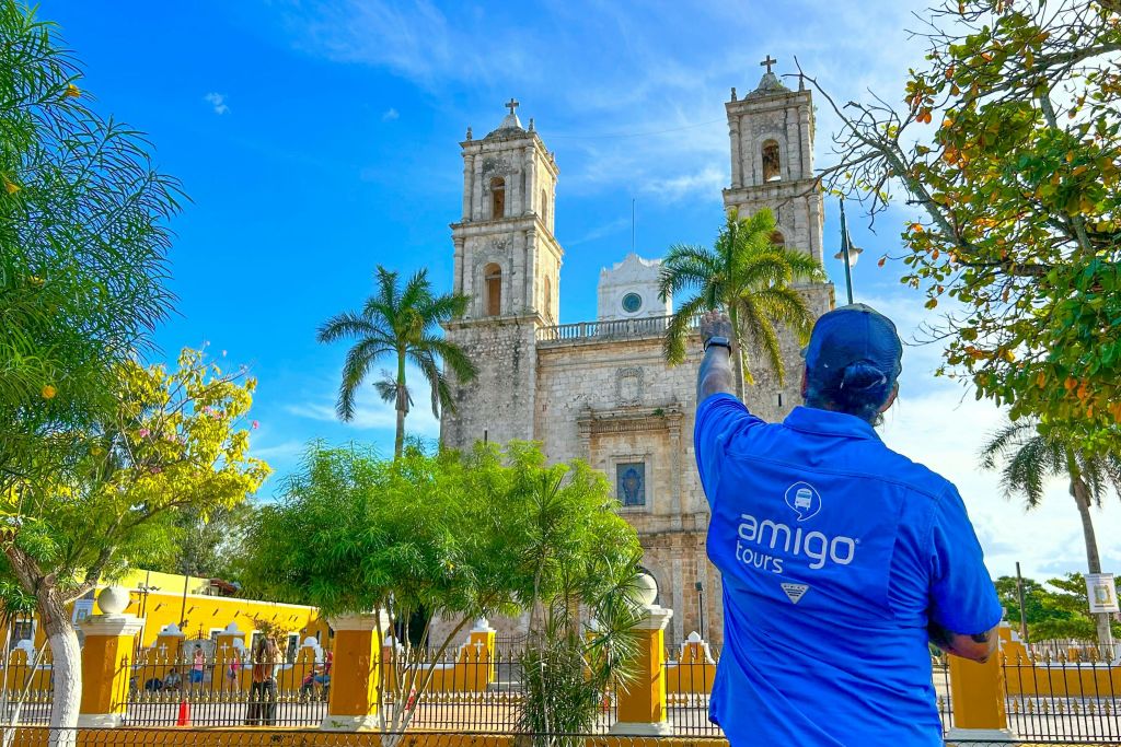 파란색 "Amigo Tours" 셔츠를 입은 사람이 나무와 푸른 하늘로 둘러싸인 두 개의 탑이 있는 유서 깊은 교회를 가리킵니다.