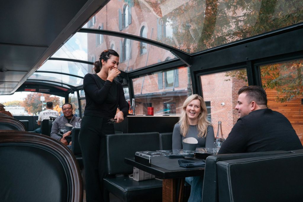 검은색 옷을 입은 여성이 유리 지붕의 버스 안에서 금발의 미소를 짓는 여성을 포함해 좌석에 앉은 승객과 함께 웃고 있습니다.