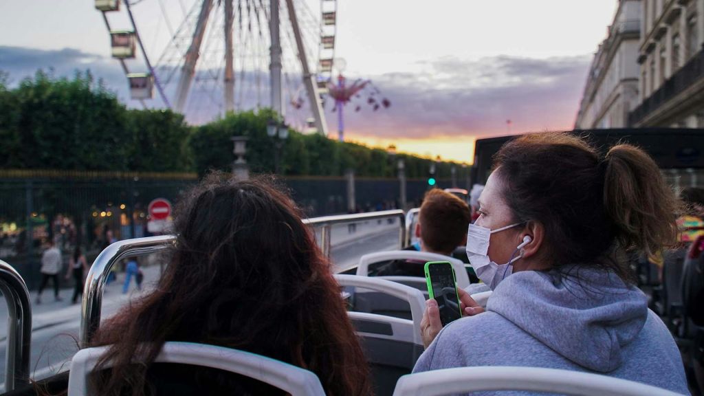 대관람차와 일몰을 배경으로 한 사람이 마스크를 쓰고 스마트폰을 보고 있는 오픈탑 버스에 탄 사람들.