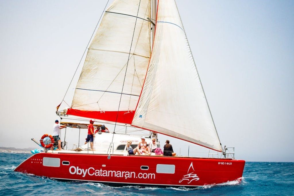 "ObyCatamaran.com" 이라는 이름의 붉은 쌍동선이 하얀 돛 아래 갑판에 있는 사람들과 함께 바다에서 항해합니다.