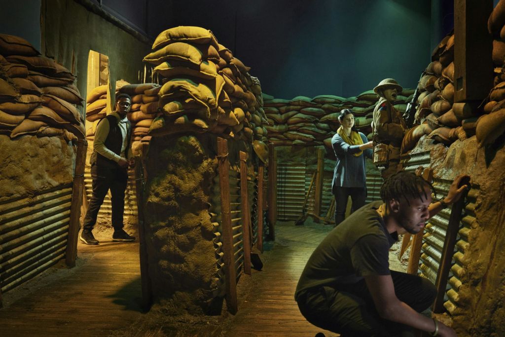 두 사람이 모래주머니와 나무 지원군이 있는 희미한 조명의 참호 전시관을 탐험하고 있습니다. 한 사람은 웅크리고 있고, 다른 한 사람은 병사 마네킹 근처에 서 있습니다.