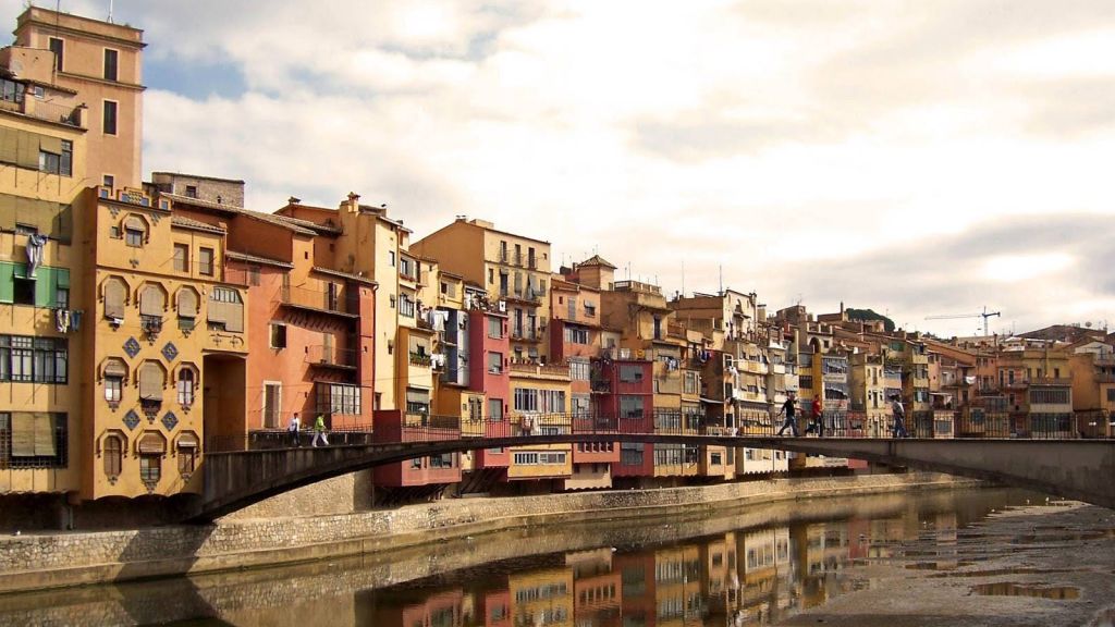 다리가 다채로운 건물 옆의 강을 가로지르며 몇 사람이 흐린 하늘 아래 다리 위를 걷고 있습니다.