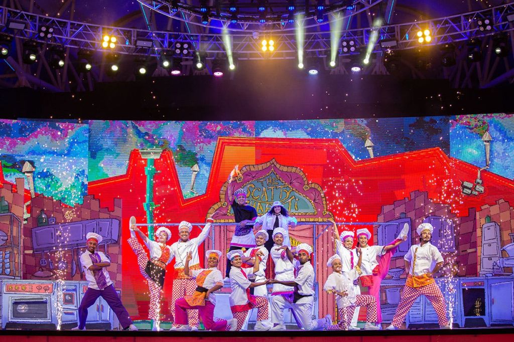 다채롭고 생동감 넘치는 주방을 배경으로 밝게 빛나는 무대에서 셰프 의상을 입은 공연자들이 춤을 추고 있습니다.