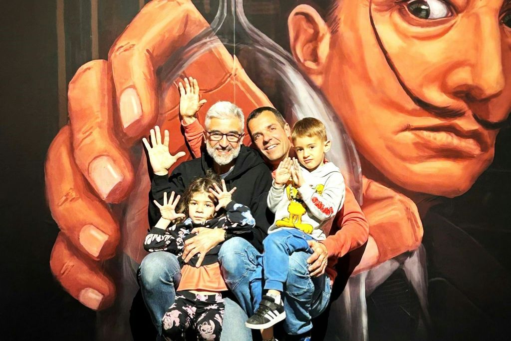 어른 두 명과 어린이 두 명이 콧수염이 달린 커다란 예술적 얼굴이 그려진 벽화 앞에서 미소를 짓고 손을 흔들고 있습니다.