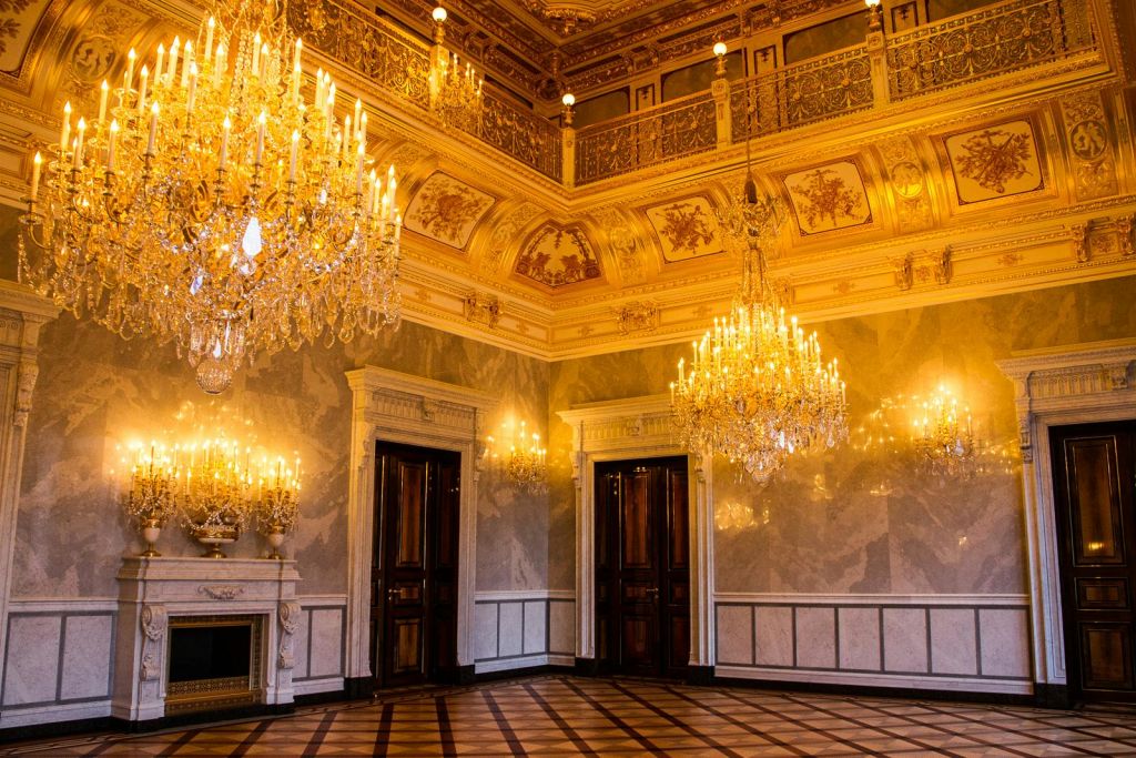 화려한 샹들리에, 황금 천장 장식, 나무 문, 체크무늬 바닥이 있는 우아한 객실입니다.