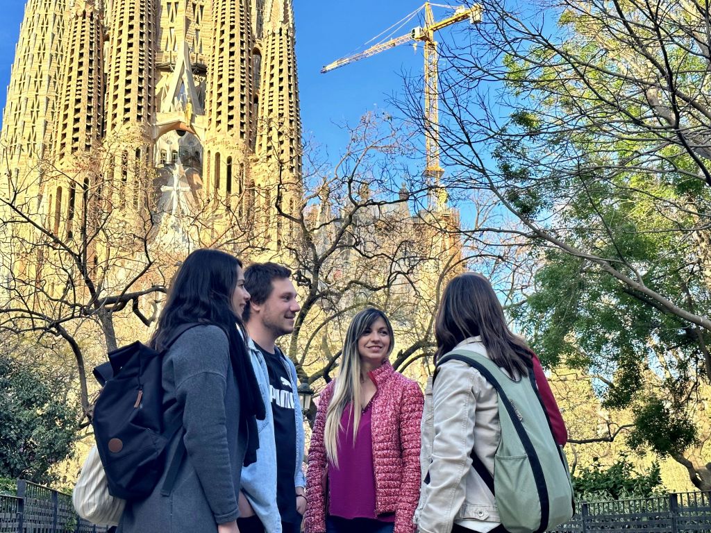 사그라다 파밀리아 (Sagrada Familia) 앞에 네 사람이 함께 서서 미소를 짓고 있습니다. 배경에는 크레인과 나무가 보입니다.