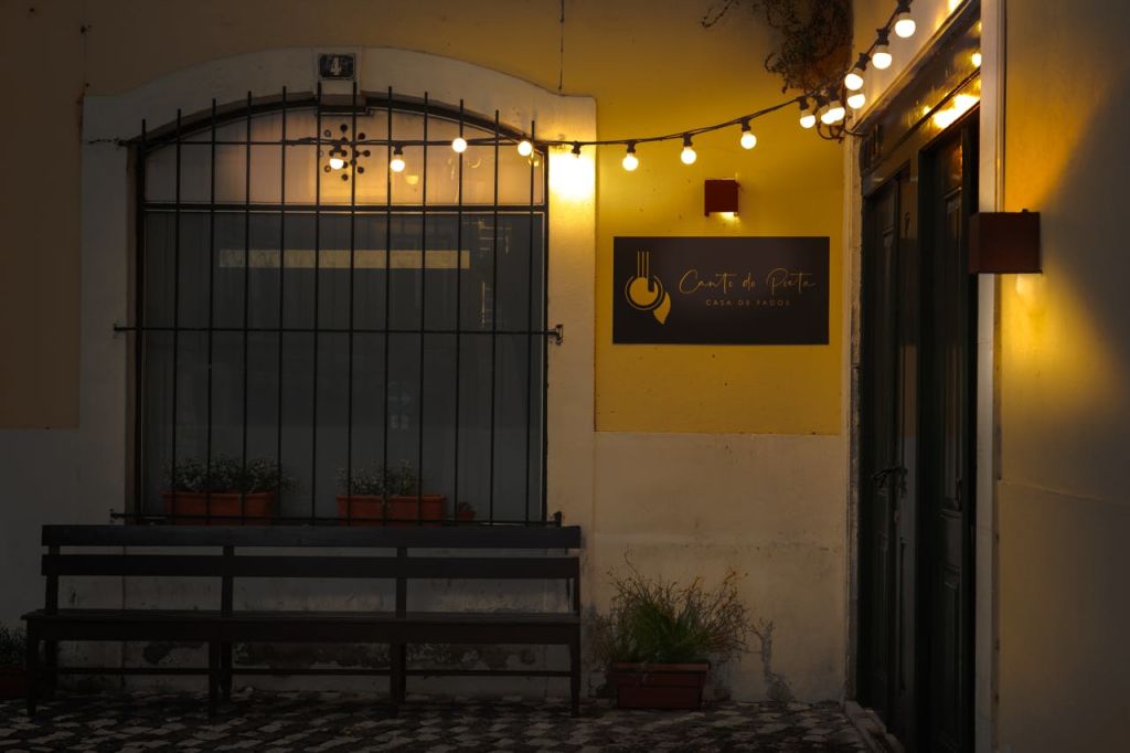 스트링 조명이 있는 야외 카페, "Canto da Peia" 표지판, 바, 화분, 벤치가 있는 창문.