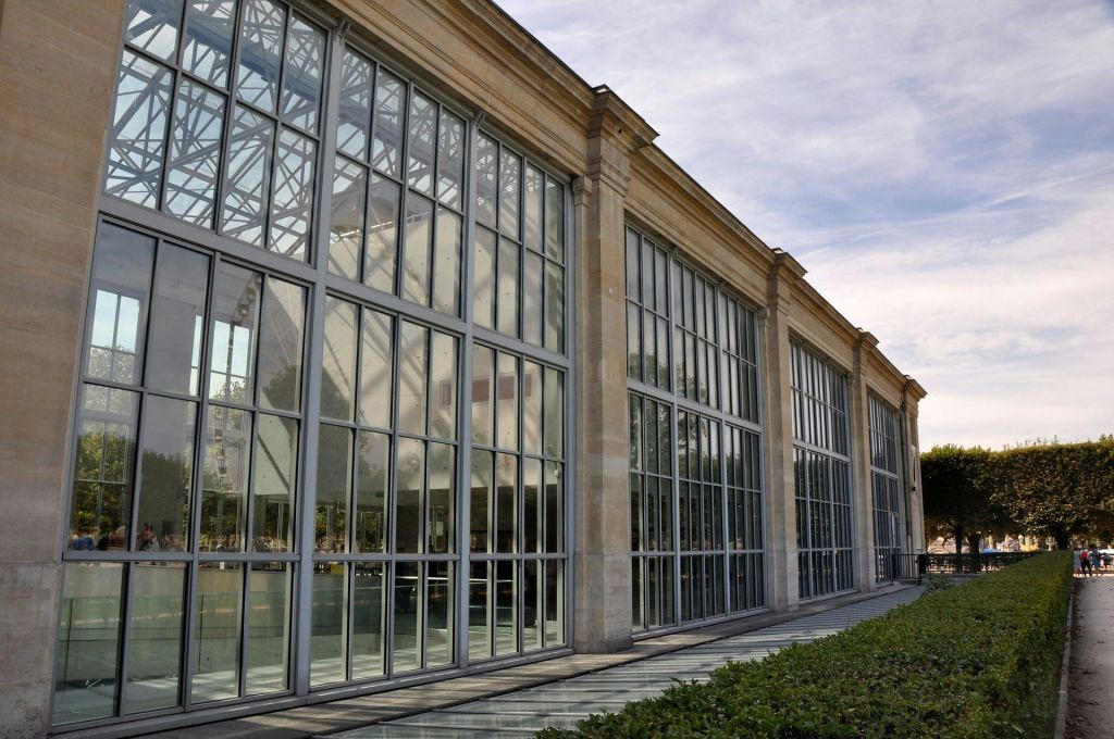 탁한 하늘 아래 깔끔하게 다듬어진 울타리 옆에 금속 프레임워크와 커다란 창문이 있는 유리로 된 건물입니다.