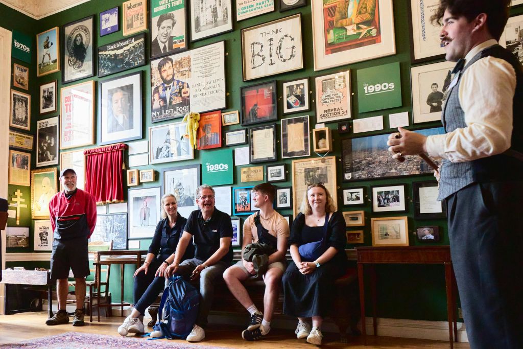 네 명이 한 방의 벤치에 앉아 여러 장의 액자에 담긴 사진, 포스터, 작품이 벽에 걸려 있습니다.