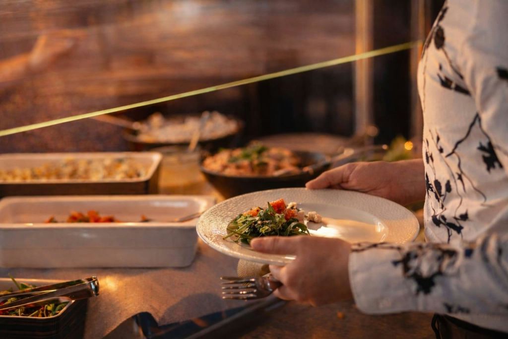 뷔페에서 샐러드가 담긴 접시를 들고 있는 사람으로, 따뜻한 조명으로 나무 테이블 위에 다양한 요리를 쟁반에 올려 놓습니다.