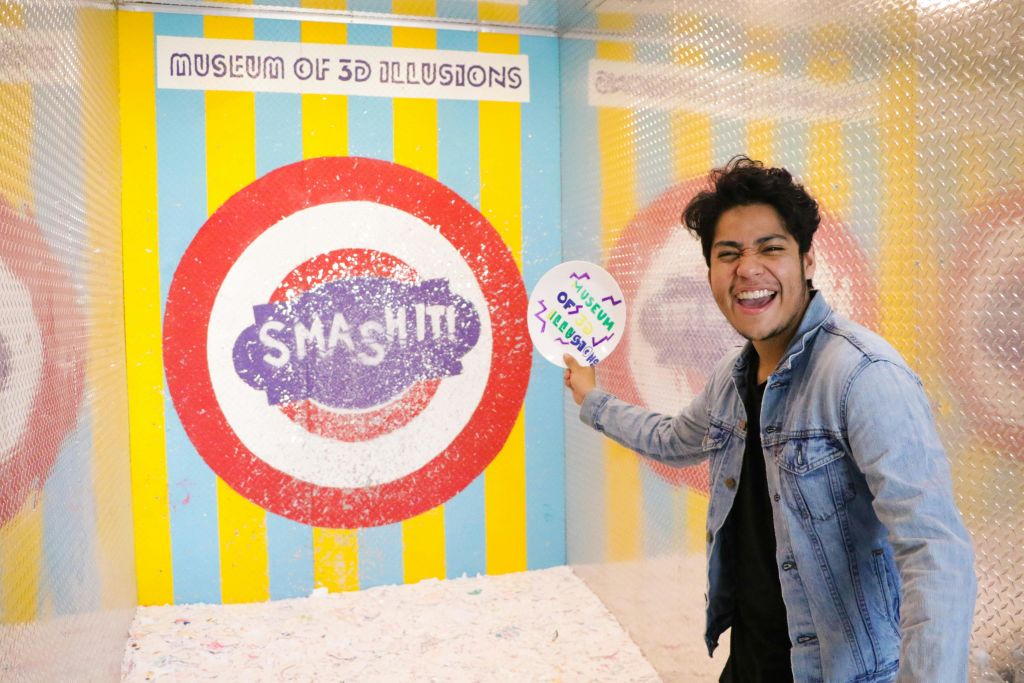 위에 쓰여진 "3D 환상 박물관" 과 다른 텍스트가 부분적으로 보이는 화려한 부스에서 웃으며 간판을 들고 있는 사람.