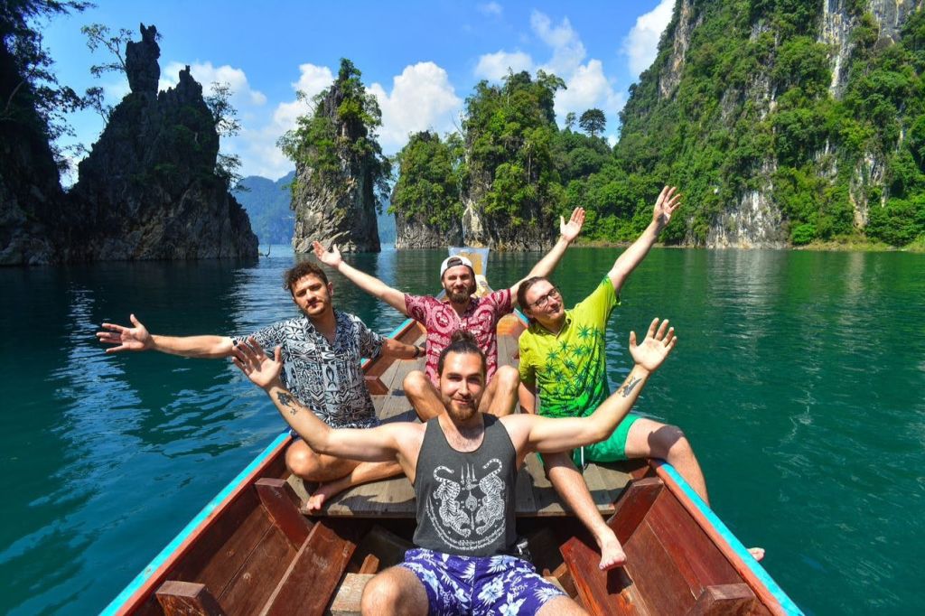 푸른 하늘 아래 높고 울창한 푸른 섬으로 둘러싸인 청록색 호수에서 팔을 뻗은 네 명의 남성이 보트에 앉아 있습니다.
