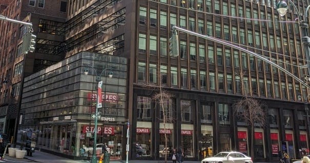 창문이 많은 건물의 거리 풍경, 1층에 스테이플스 매장, 앞쪽으로 지나가는 자동차.