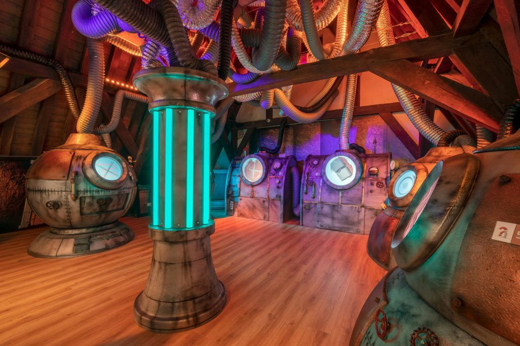 금속 포드, 원형 문, 눈에 띄는 파이프, 빛나는 파란색 기둥이 있는 미래형 객실입니다. 나무 바닥이 인더스트리얼 인테리어와 대조를 이룹니다.